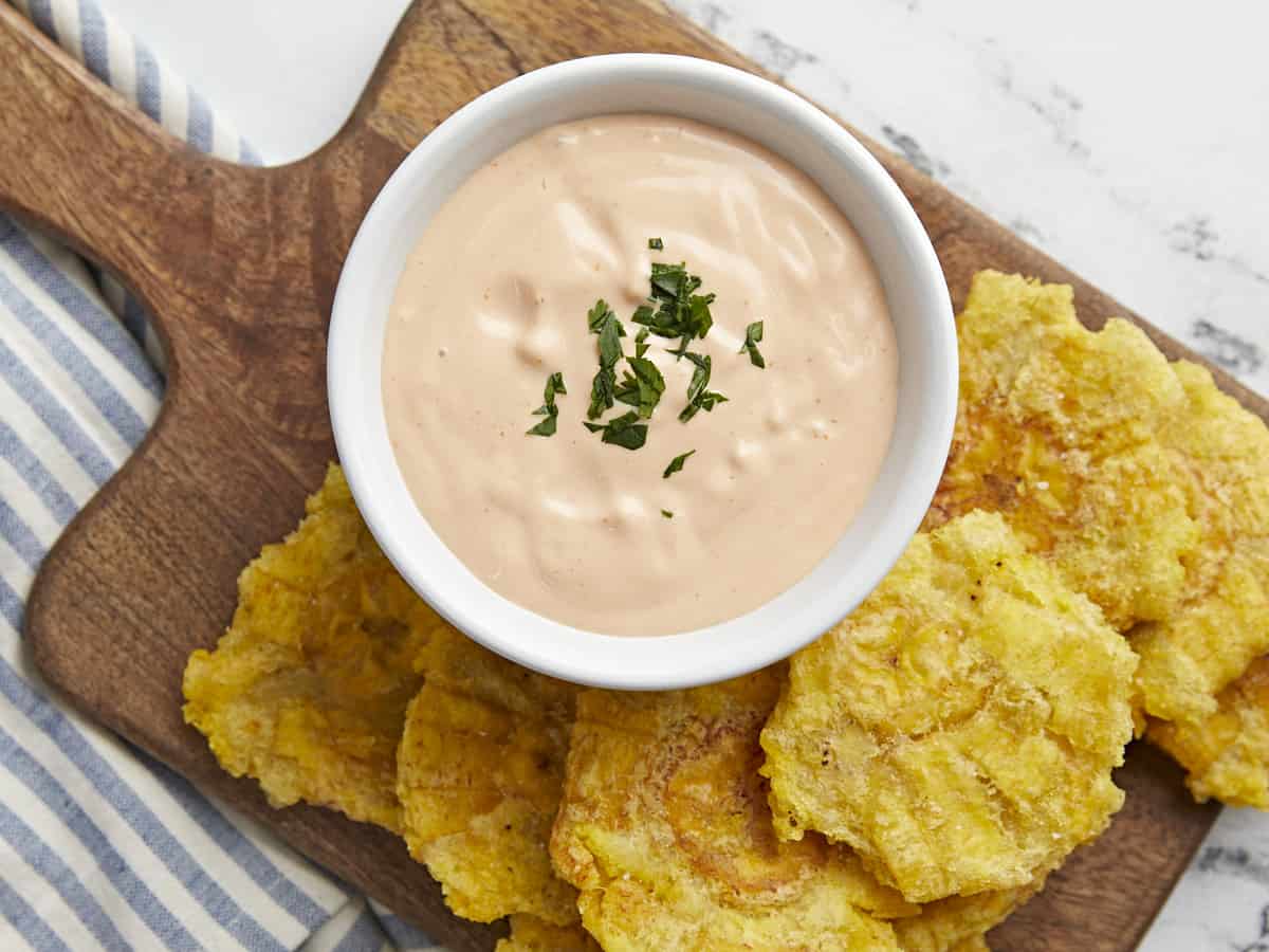 A top-down view of creamy mayo-ketchup farmacias savia, sauce in a white bowl, garnished with chopped green herbs, served with golden yellow fried plantain chips on a wooden serving board