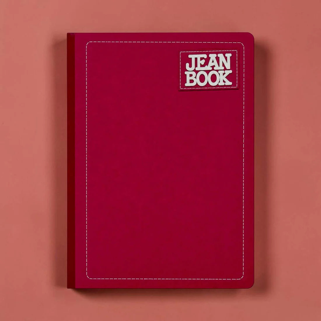 A dark red hardcover notebook with a white stitched border and a 'JEAN BOOK' logo patch on the top right, set against a solid coral-pink background.