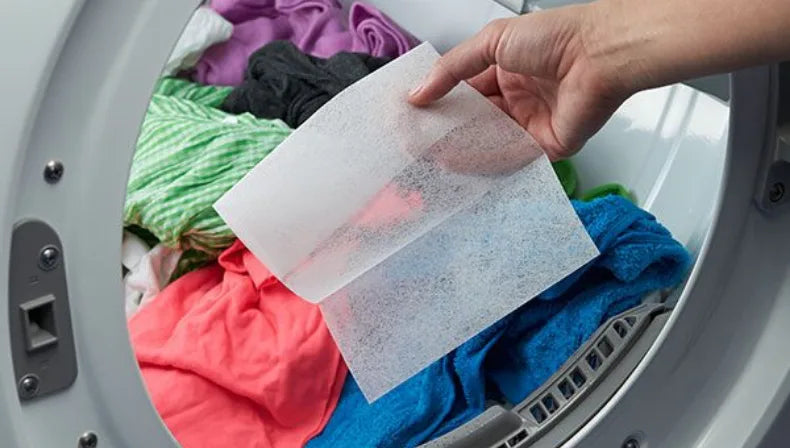 A hand holds a translucent white mesh filter sheet over the open door of a washing machine filled with colorful laundry.