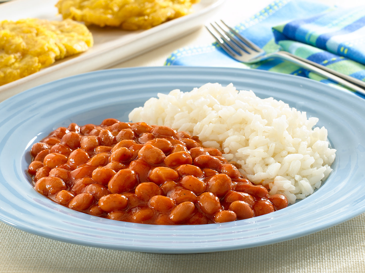 Blue plate with baked beans and white rice on a table with cornbread in the background.