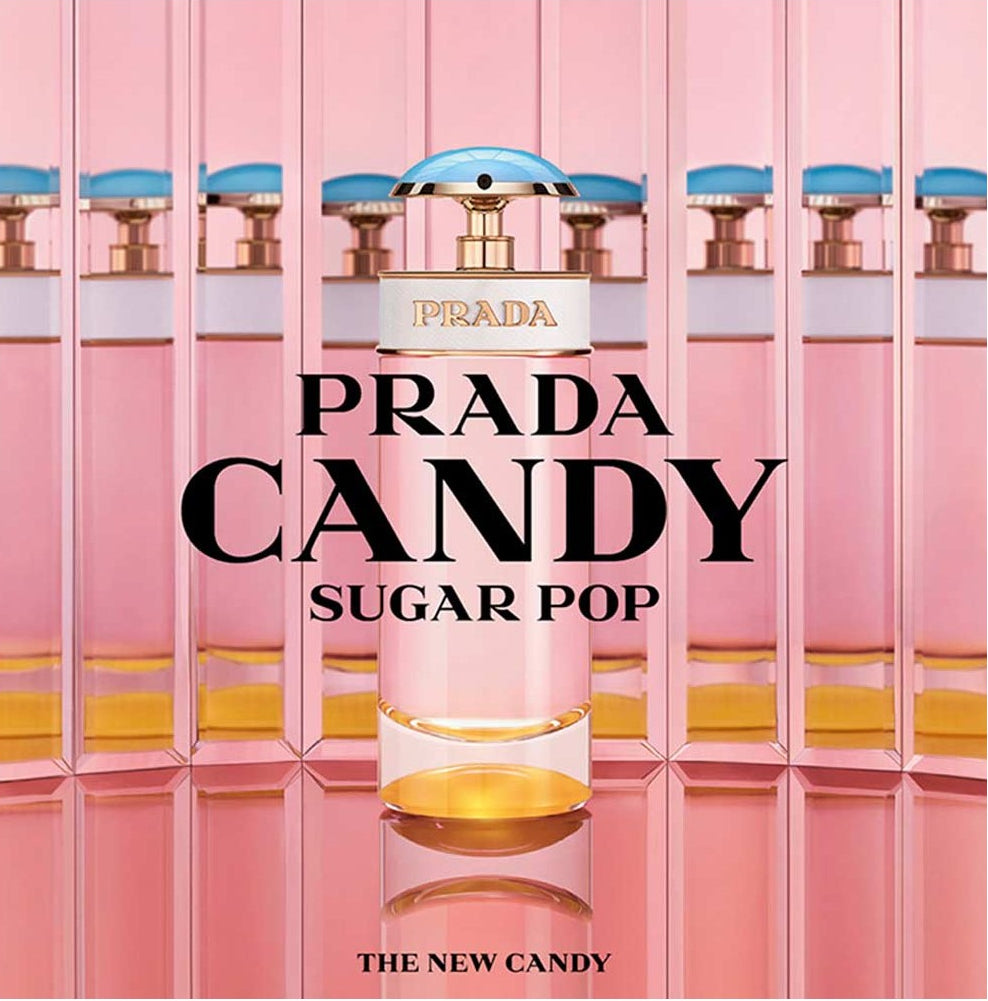 Prada Candy Sugar Pop perfume bottle centered on a pink reflective background with repeated bottle silhouettes behind it. The Prada logo is visible on the label, and large text reads 'PRADA CANDY SUGAR POP' with 'THE NEW CANDY' near the bottom.